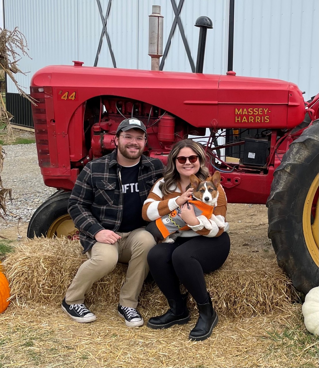 two people sitting with a dog on a hay bale in front of a tractor